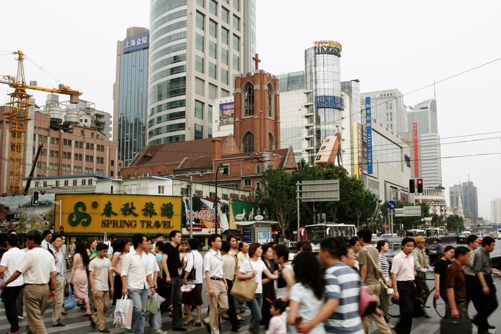 Menschen überqueren auf einem Zebrastreifen eine Straße am Platz des Volkes in Shanghai. Im Hintergrund sieht man eine christliche Kirche vor Hochäusern aus Glas.