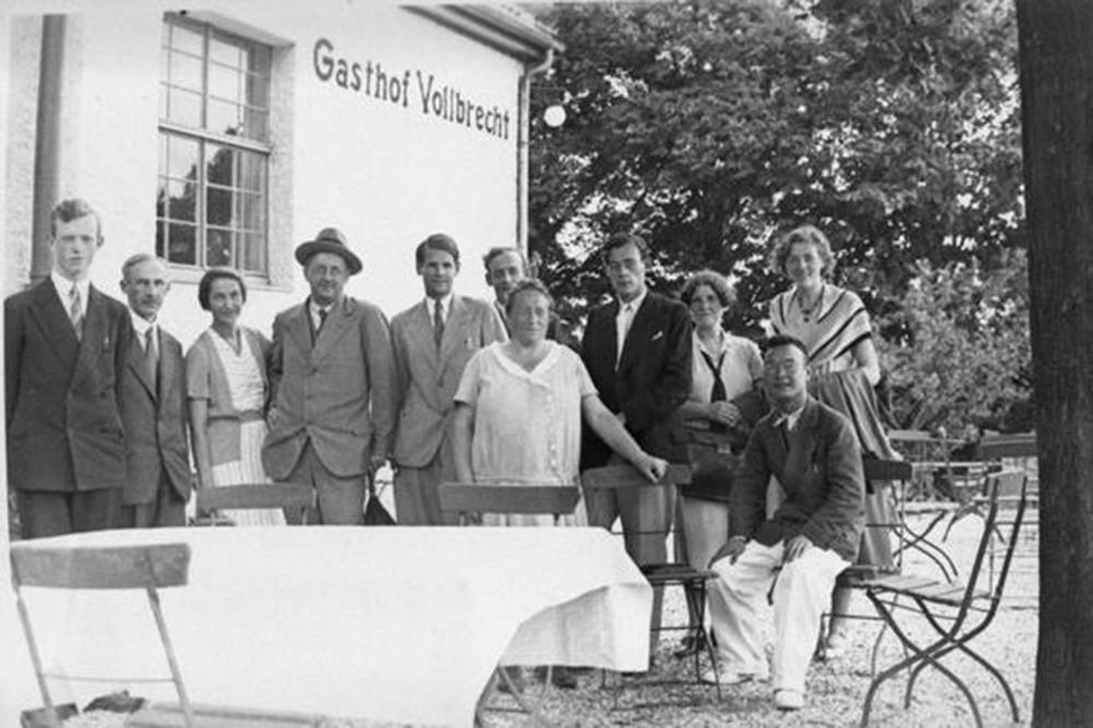 Schwarz-weiß Foto. Die Gruppe um emmy Noether hat sich zum Gruppenbild aufgestellt. Es ist Sommer, sie stehen vor einem Gasthof zwischen Tischen und Stühlen. Es ist Sommer.